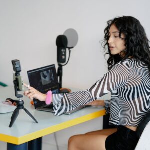 A woman recording herself with a smartphone and laptop at a desk indoors.